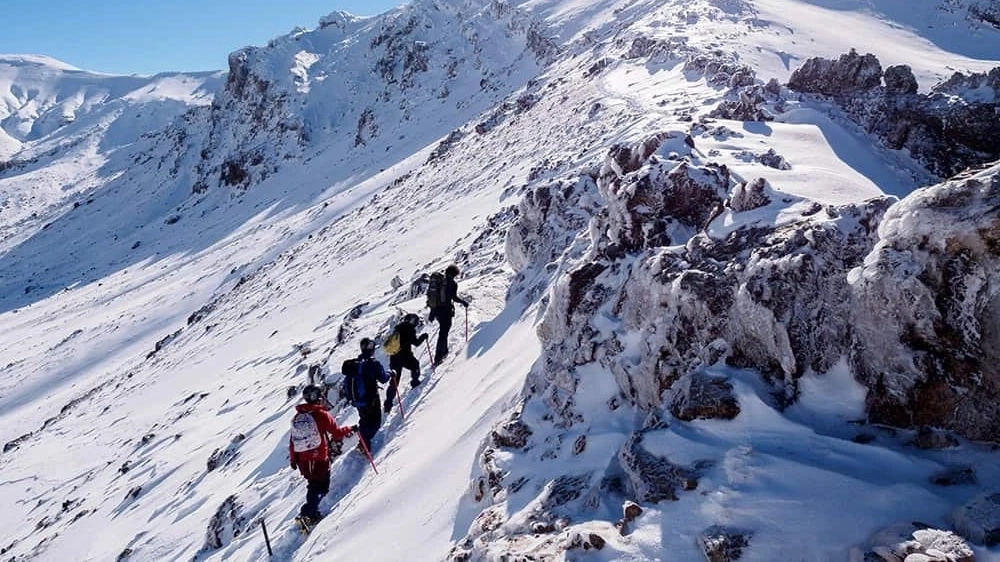 Four people in climbing gear walking up the side of a snow-covered mountain.