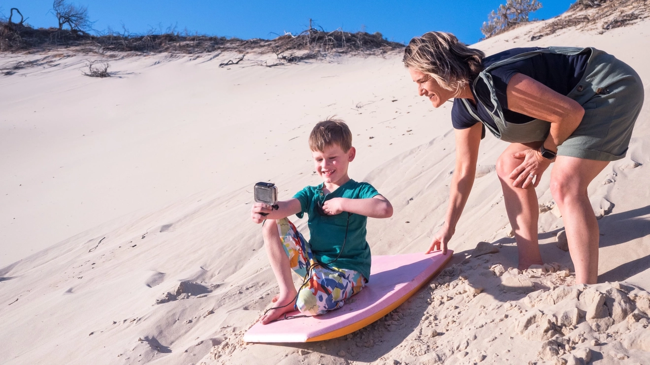 Mother helping her son on a toboggan while he takes a photo, at the sandblow on K'gari (Fraser Island). Image credit: Tourism and Events Queensland