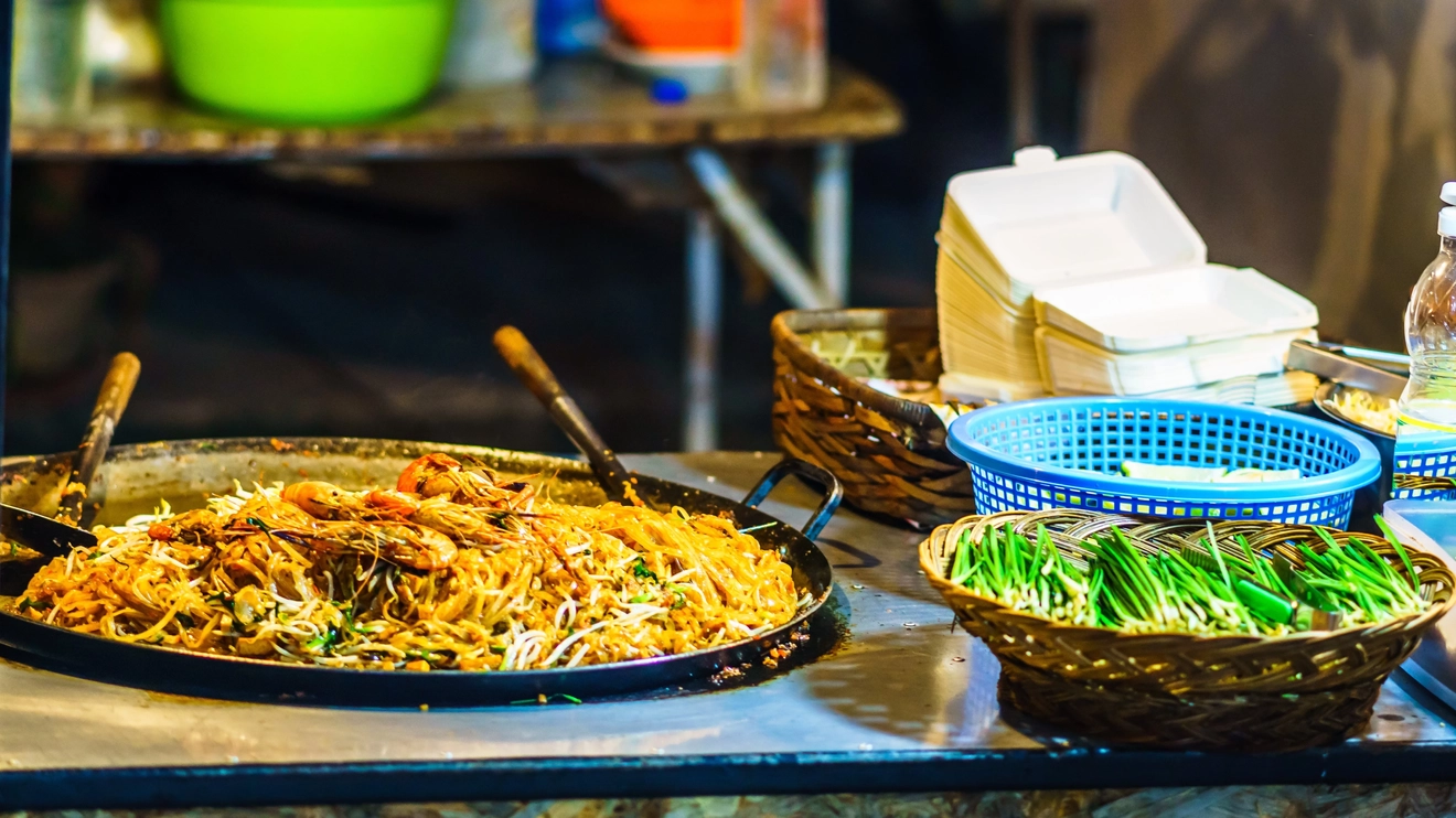Close up of a street-food stall with a wok full of noodles and prawns, and a plastic basket of greens, in Bangkok, Thailand. Image credit: stock.adobe.com