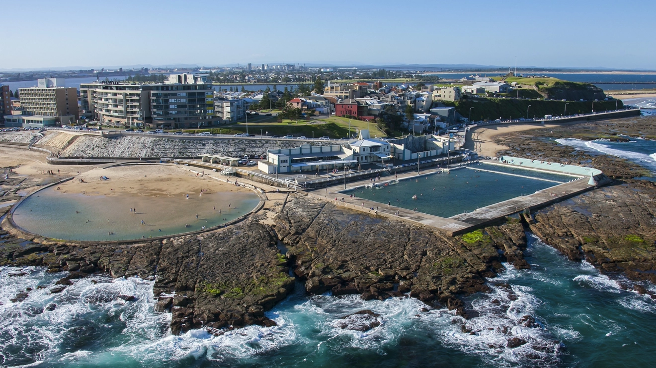 Aerial view of Newcastle Ocean Baths and Canoe Pool with city skyline in background. Image credit: Destination NSW