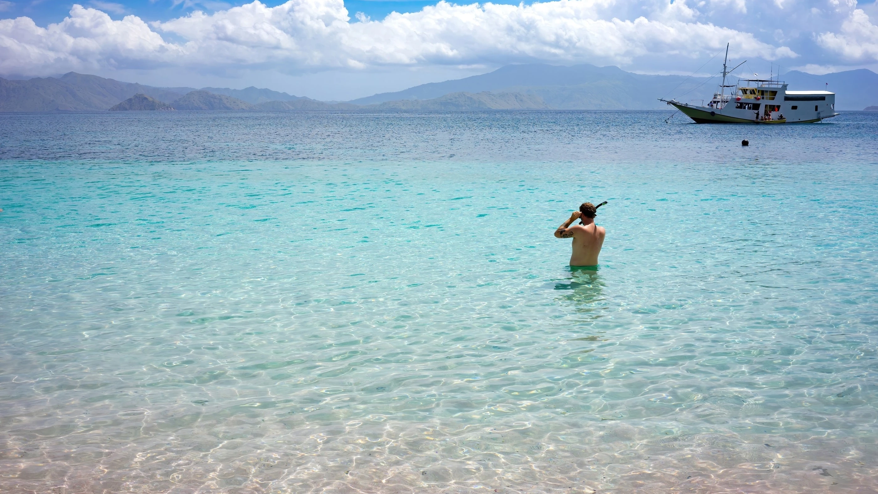 Man about to go snorkelling in the calm blue sea of Komodo National Park. A ‘phinisi’ floats nearby, and there are mountains on the horizon. Image credit: stock.adobe.com