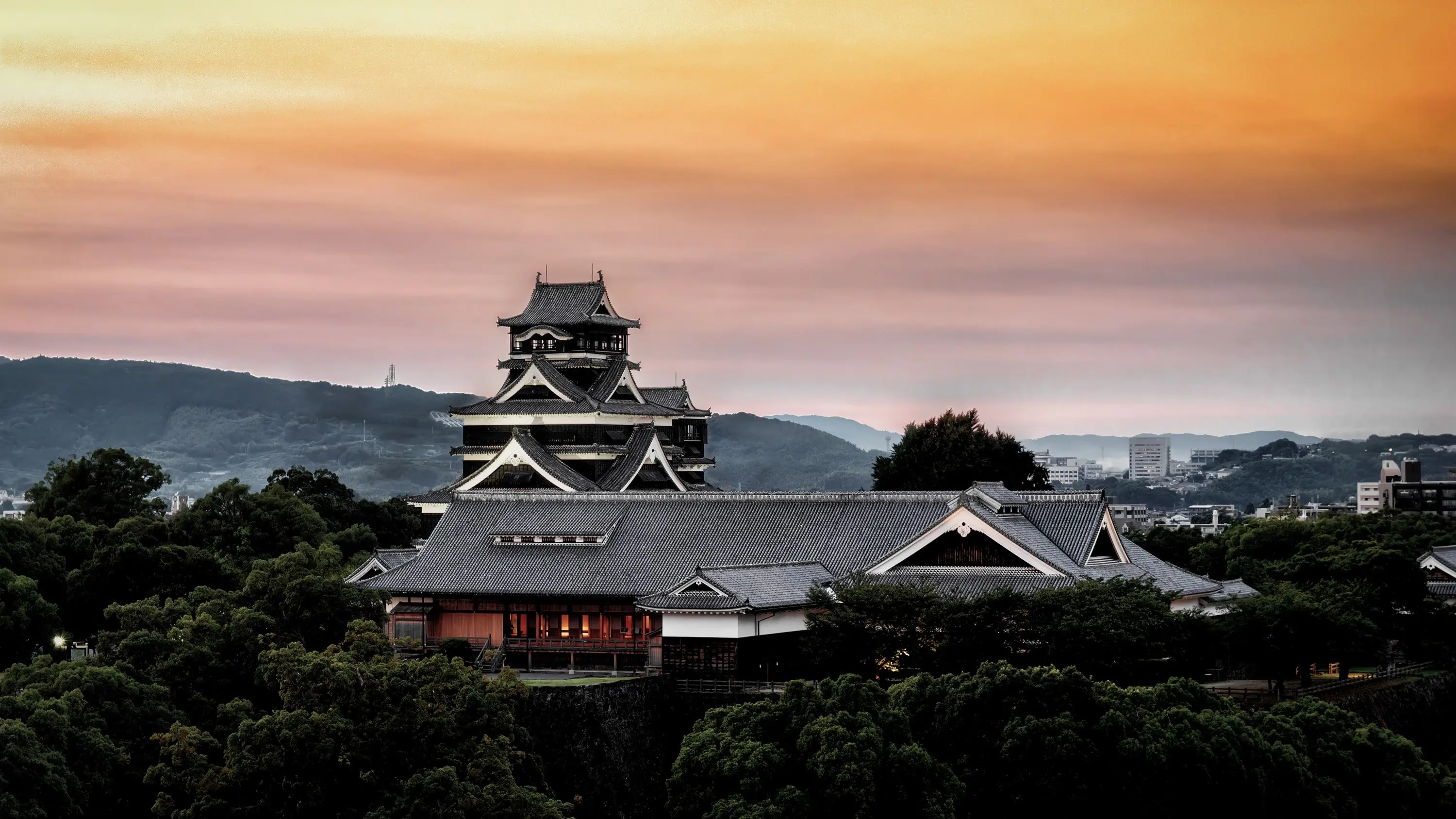 Kumamoto Castle, surrounded by trees, with Kumamoto city skyline and mountains in backround, at sunset. Kumamoto, Kyushu, Japan. Image credit: stock.adobe.com