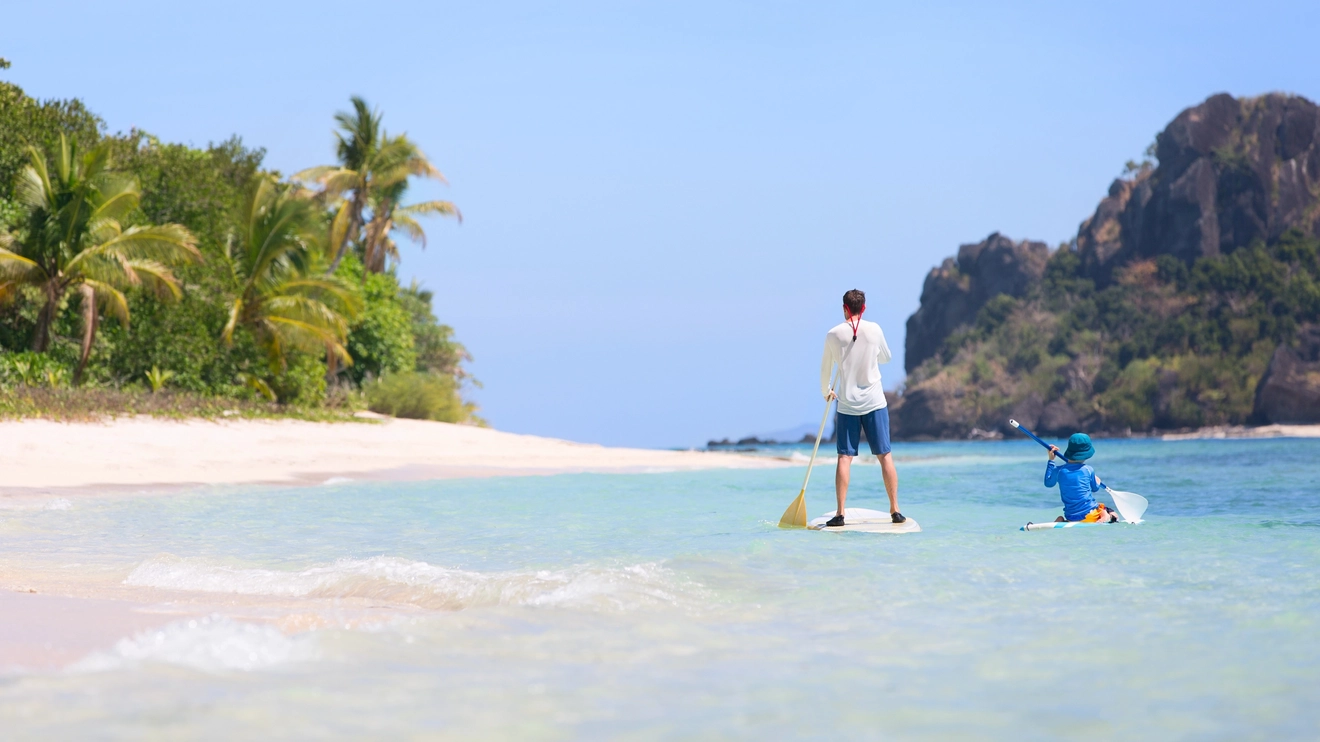 Back view of a man standing on a paddle board in the water at a white sand beach in Fiji, with a child sitting on one next to him. Image credit: stock.adobe.com