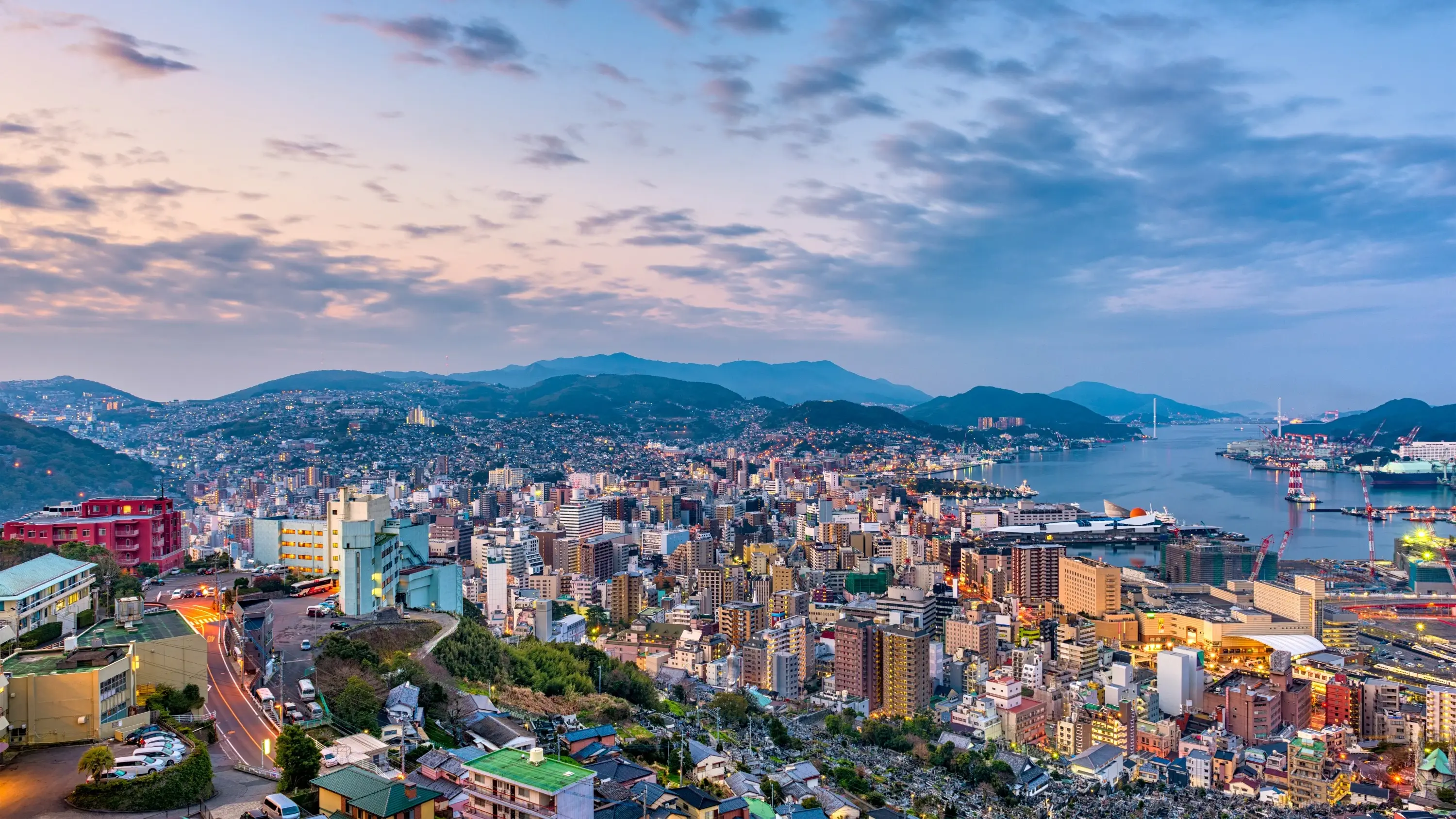 Aerial view of Nagasaki's skyline at dusk, with tall buildings in the foreground, a river and mountains in the background. Image credit: stock.adobe.com