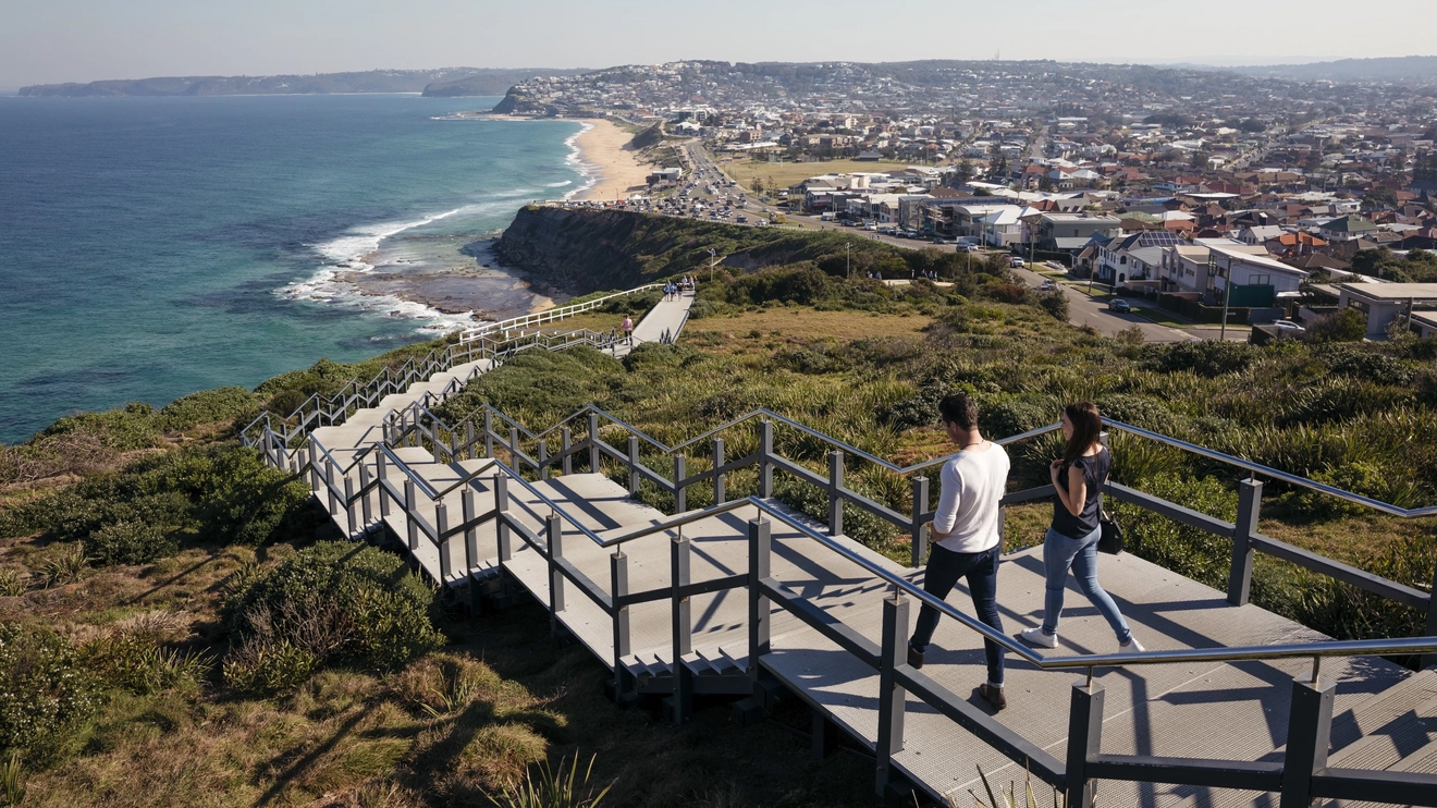 Two people descending the stairs on Newcastle Memorial Walk, with coastal and city skyline views in the background. Image credit: Destination NSW