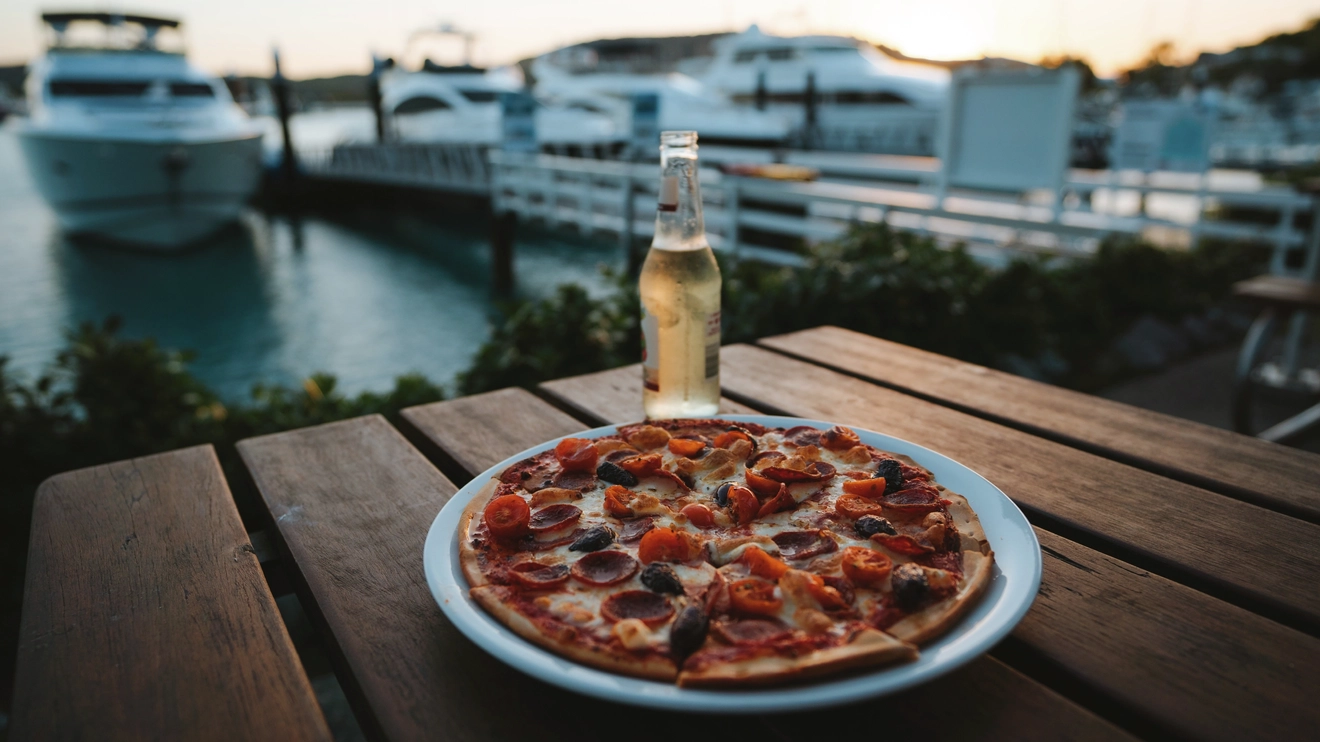 Close-up of a pizza and beer, with marina in the background, at Pizzeria and Gelato Bar, Hamilton Island. Image credit: Tourism and Events Queensland