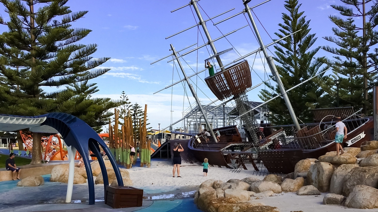 Children's shipwreck playground on the Busselton foreshore with tall pine trees and blue afternoon sky in background. Image credit: MPH Photography/Shutterstock.com
