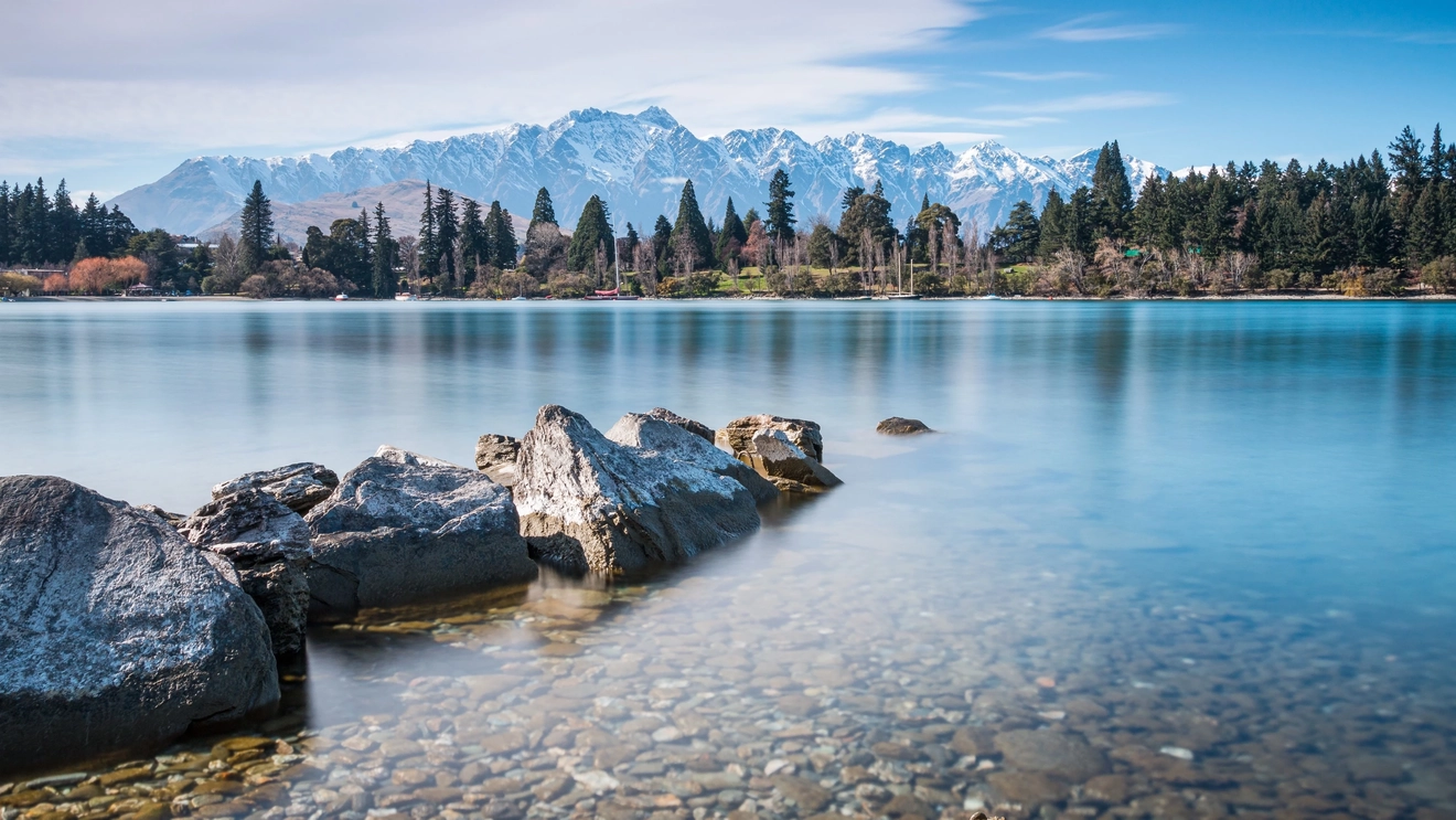 Rocks and clear water at Lake Wakatipu surrounded by trees, the Remarkables in the background, Queenstown, New Zealand. Image credit: stock.adobe.com